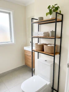 Slim black metal etagere above a white toilet with bamboo shelves, folded towels, woven baskets, and spare toilet paper.