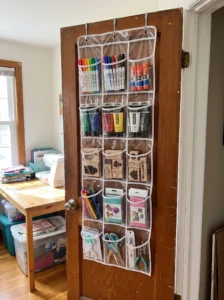Clear over-the-door organizer storing markers, glue sticks, paint tubes, stamps, and small craft supplies.