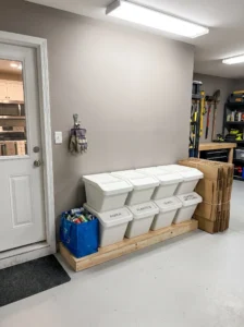 Organized garage recycling station with labeled sorting bins, a reusable bag for bottles and cans, and flattened cardboard.